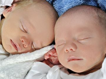 Sie sehen Zwillinge im Säuglinsalter. Beide Kinder schlafen und tragen einen weißen Schlafanzug
